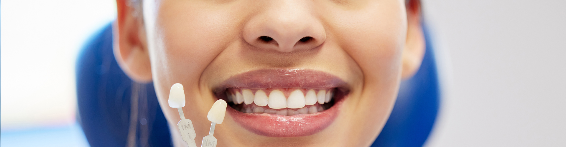 Gloved hand holding tooth shade-matching tool next to woman's smile.